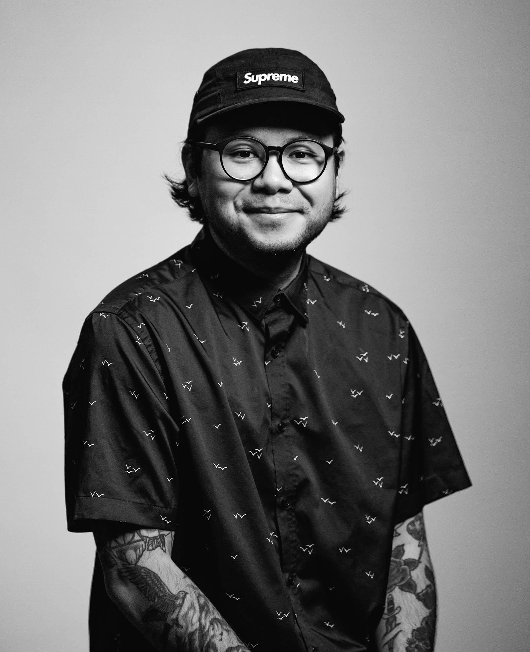 Antonio Phumipraphat, content creator for Think3r Networks a part of Think 3D Solutions, half smiling with black hat with white writing, longer flipping out dark hair seen sticking out the sides of hat, dark-rimmed glasses, subtle facial hair, dark button-up shirt with white 'V' shapes on it, medium skin complexion, arms seen with tattoos, the photo is in black and white.