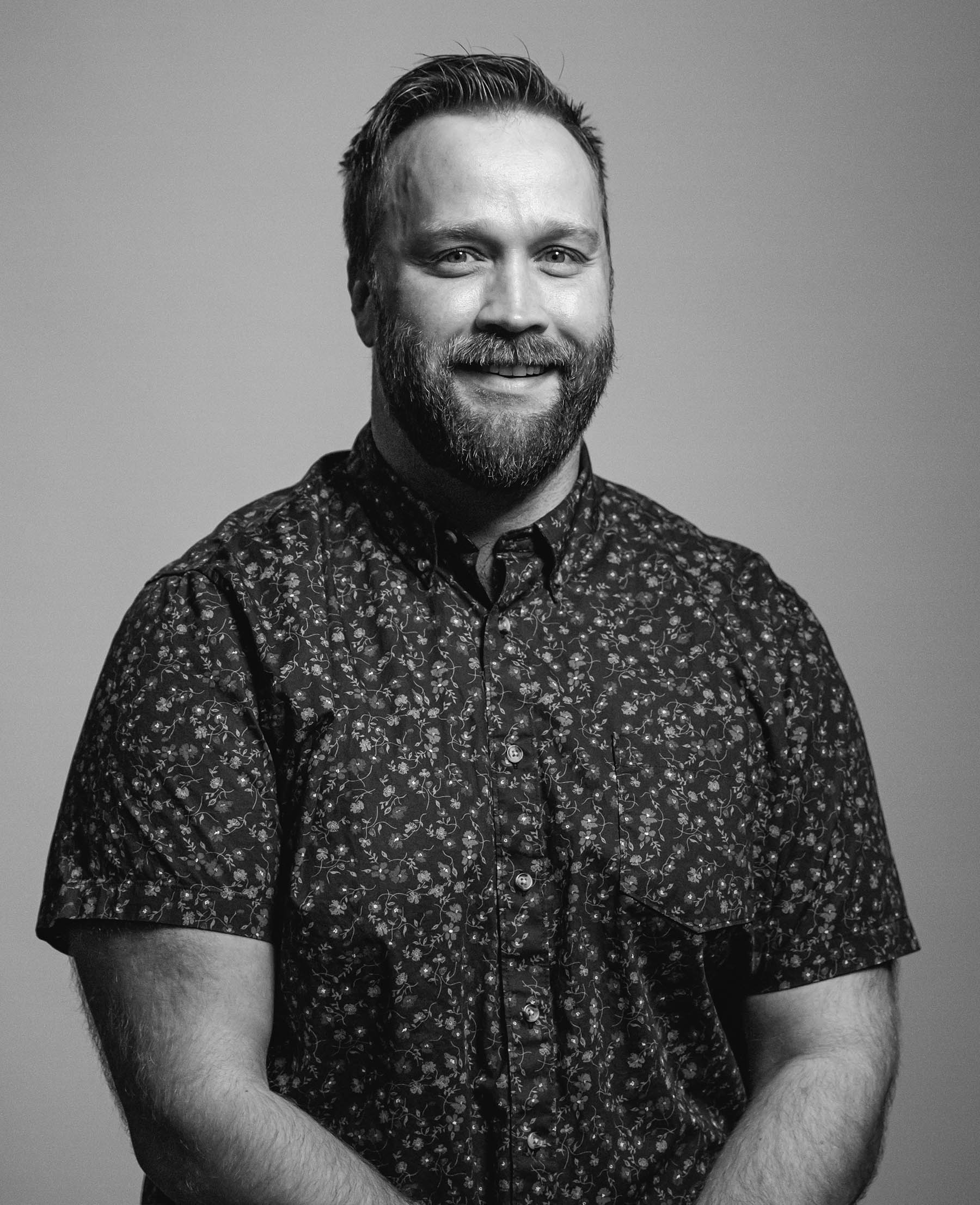 Brett Feterl, director of community relations for Think 3D Solutions, shorter medium colored hair, full beard, button-up shirt with light and dark pattern, lighter complexion, the photo is in black and white.