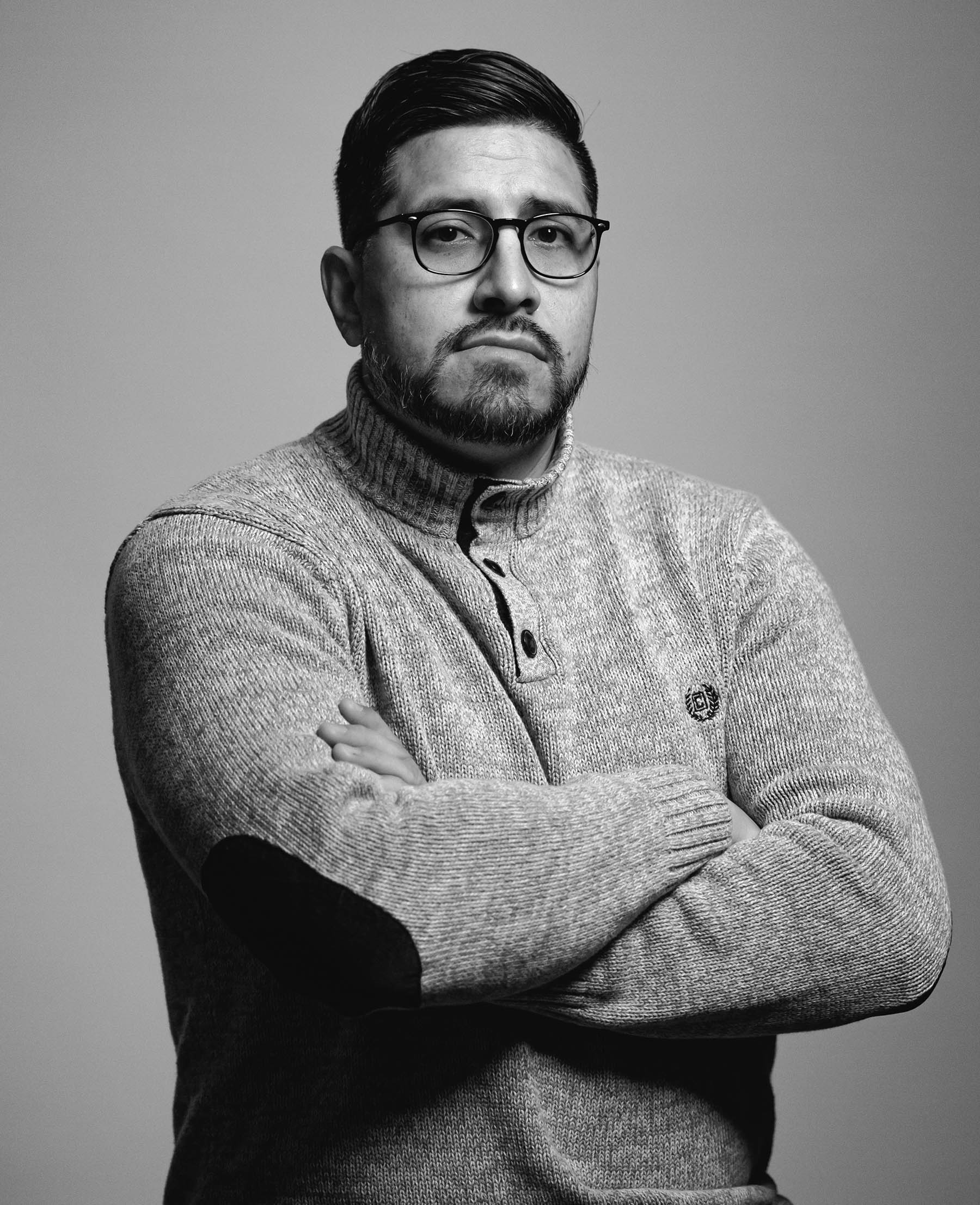 Jesus Gonzalez, videographer and community coordinator for Think 3D Solutions, stoic face, shorter dark hair brushed back, beard, dark rimmed glasses, arms crossed across chest, lighter sweater with 3 buttons, medium complexion, the photo is in black and white.