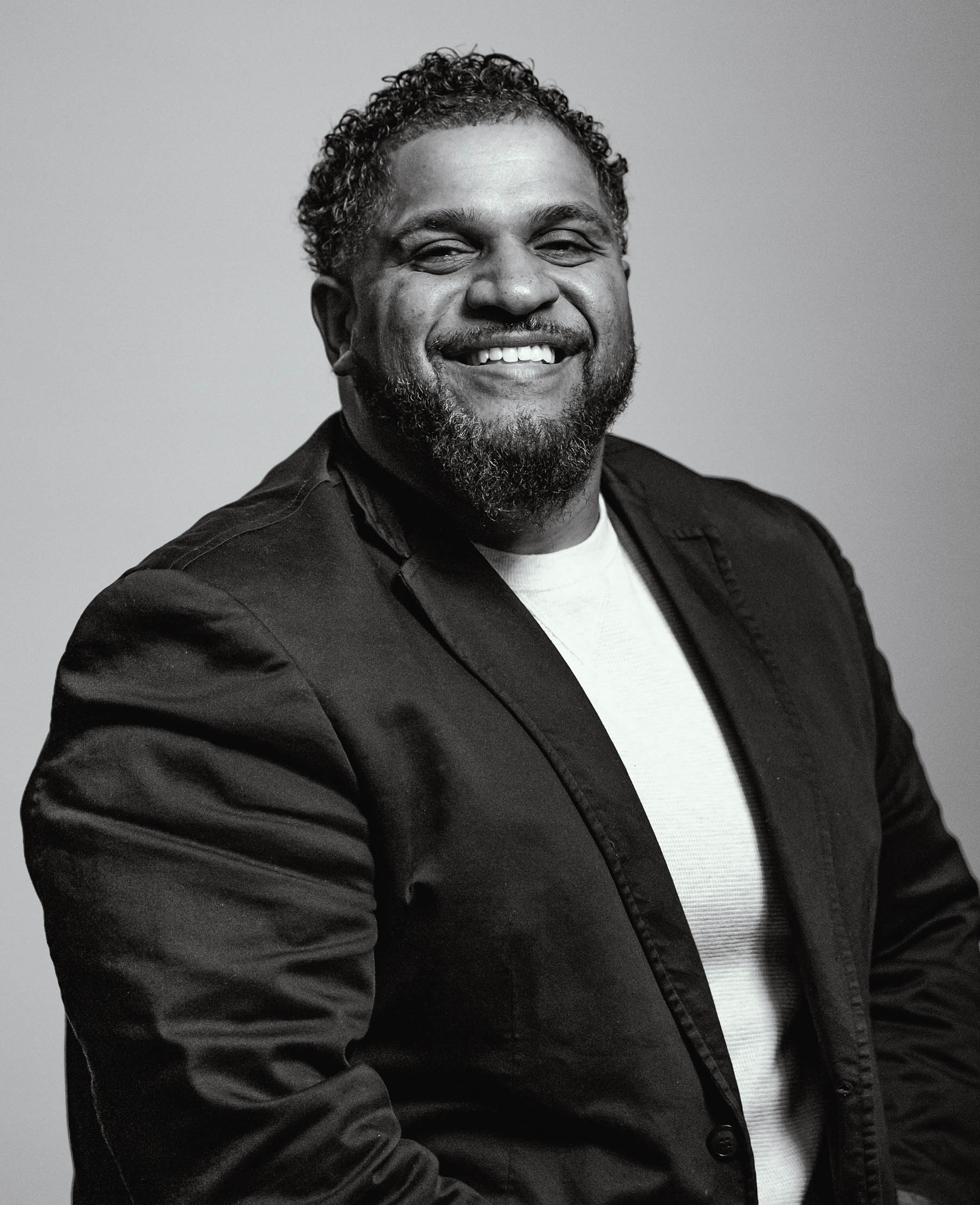 Manny Gutierrez, director of community relations for Think 3D Solutions, has dark curly hair, a broad smile, a beard, a black blazer with a white shirt underneath, and wide shoulders, the photo is in black and white.