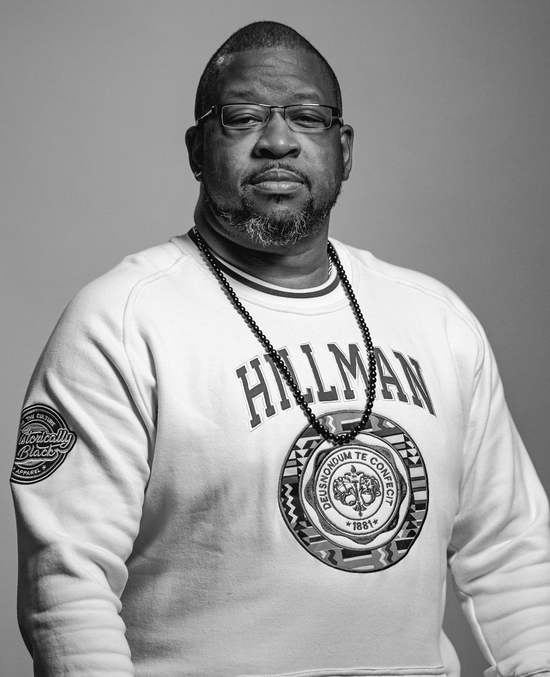 Vaney Hariri, chief heart officer and co-founder of Think 3D Solutions, is sitting with a stoic look on his face, glasses, close-cut dark hair, dark completion, beard, white Hillman sweatshirt, and necklace, photo is in black and white.